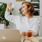 Smiling woman holding cash, working at home office with a laptop, symbolizing financial success.